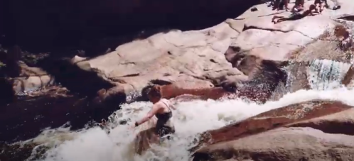 A person wades through a rocky stream with cascading water, while others relax on the rocks above.