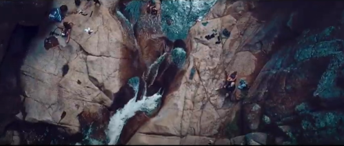 Aerial view of a rocky riverbed with people relaxing by the water and a small waterfall flowing through the rocks.