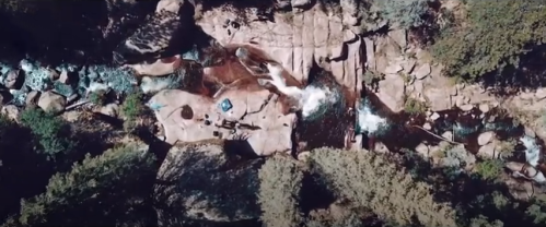 Aerial view of a rocky riverbed with flowing water, surrounded by trees and large boulders.