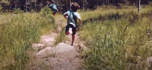 A child runs along a dirt path through tall grass and trees, enjoying a sunny day outdoors.