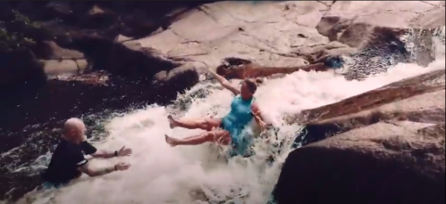 A person in a blue dress slides down a rocky waterfall while another person reaches out to help.