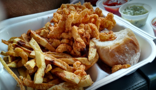 A takeout container filled with crispy fried shrimp, golden fries, and a soft roll, accompanied by various dipping sauces.