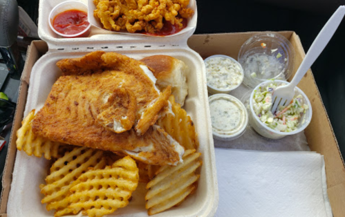A takeout box with fried fish, waffle fries, coleslaw, and dipping sauces.
