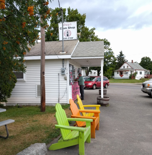 A small ice cream shop with colorful chairs outside, located on a quiet street with a few cars parked nearby.