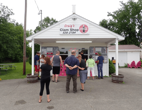 A group of people waiting in line outside Craig's Clam Shop, a small building with a menu board.