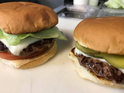 Two burgers on a white surface, one with lettuce and tomato, the other with pickles and mayonnaise.