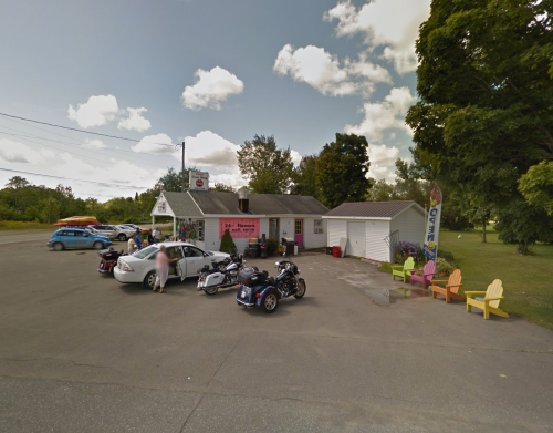 A small roadside shop with colorful chairs outside, parked cars, and motorcycles in a rural setting under a cloudy sky.