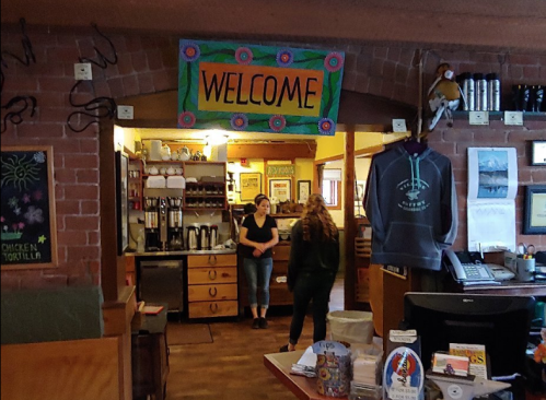 A cozy café interior with a colorful "WELCOME" sign, featuring a counter, coffee station, and two people chatting.