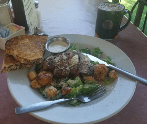 A plate with sliced meat, salad, croutons, and a side of dressing, accompanied by toast and a coffee mug.