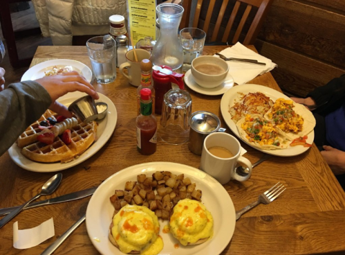 A table set with breakfast dishes: waffles with fruit, eggs Benedict, hash browns, and coffee.