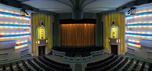 Art Deco theater interior with colorful walls, a stage draped in gold curtains, and rows of empty red seats.