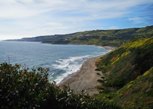 A scenic coastal view with waves lapping a sandy beach, surrounded by lush green hills under a blue sky.