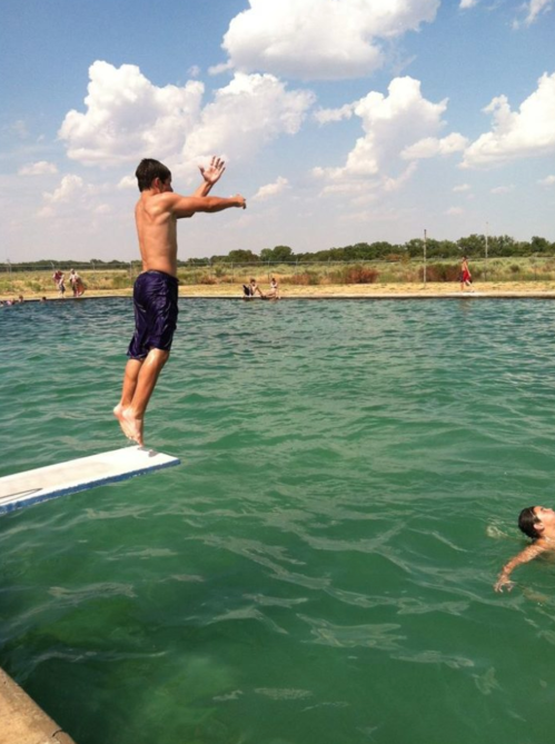A shirtless boy jumps off a diving board into a green lake, with people swimming and enjoying the sunny day.