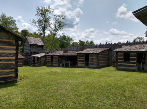 A historic settlement with log cabins and a wooden fence, set in a grassy area under a partly cloudy sky.