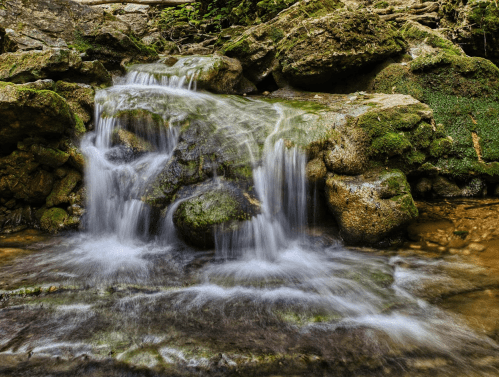 A serene waterfall cascading over moss-covered rocks in a lush, green forest setting.