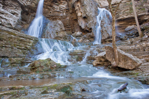 A serene waterfall cascading over rocky cliffs into a tranquil pool, surrounded by trees and natural stone formations.