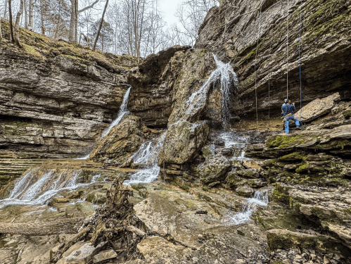 A person rappels down a rocky cliff beside cascading waterfalls in a forested area.