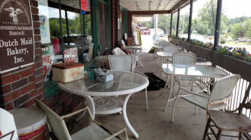 A porch with empty tables and chairs outside a bakery, surrounded by flowers and a view of a parking lot.