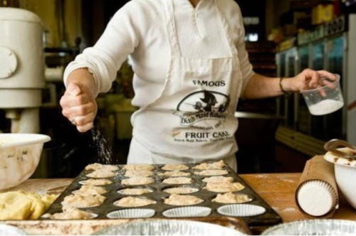 A baker sprinkles sugar over muffin batter in a kitchen, with trays of unbaked muffins ready for the oven.