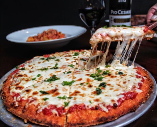 A cheesy pizza with a golden crust, topped with marinara sauce and herbs, being served with a glass of wine in the background.