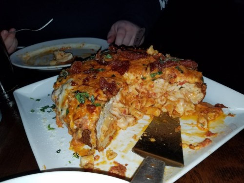 A slice of cheesy lasagna topped with bacon and herbs on a white plate, with a fork and empty plate in the background.