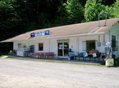 A small, rustic restaurant with a sign, outdoor seating, and a Pepsi advertisement, surrounded by greenery.