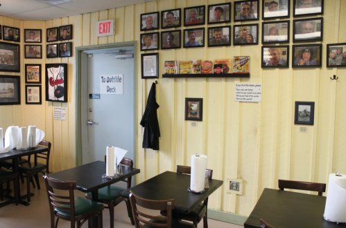 Interior of a restaurant with wooden walls, tables, and a wall of framed photos. Exit door leads to an outside deck.