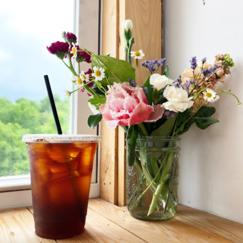A glass of iced coffee sits next to a vase of colorful flowers on a wooden table by a window.