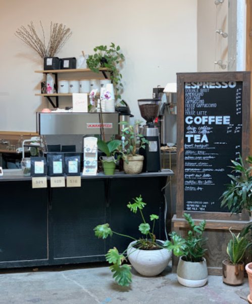 A cozy coffee shop counter with a menu board, espresso machine, and plants in decorative pots.