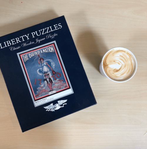 A box of Liberty Puzzles titled "The Birth of a Nation" next to a cup of latte art on a wooden table.