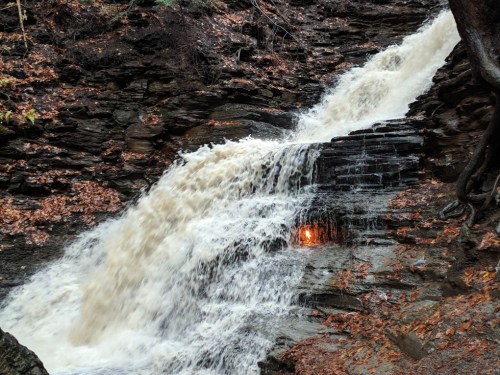A cascading waterfall flows over rocky terrain, surrounded by autumn leaves and a hint of sunlight peeking through.
