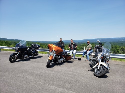 Three people stand beside their motorcycles on a scenic overlook with a clear blue sky and green hills in the background.