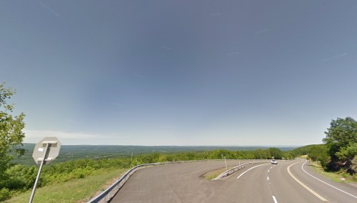 A winding road curves through a green landscape under a clear blue sky, with distant hills visible in the background.