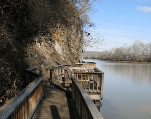 A wooden walkway curves along a river, flanked by rocky cliffs and bare trees under a clear blue sky.