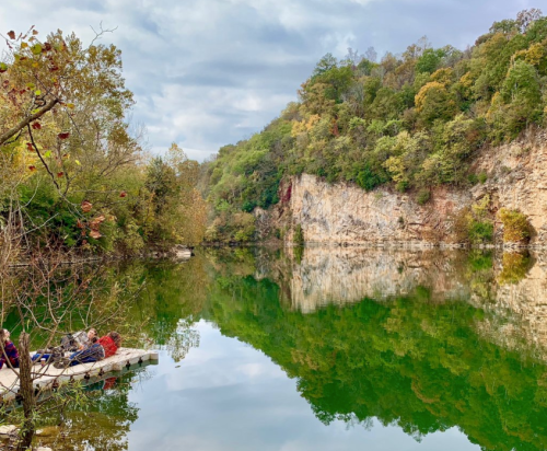A serene lake surrounded by trees and cliffs, reflecting the landscape in calm waters, with people sitting on a dock.