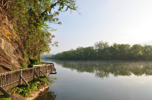 A serene river scene with a wooden walkway along a rocky bank, surrounded by lush greenery and morning mist.