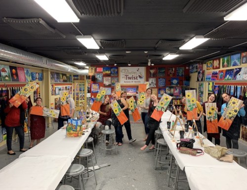 A group of people holding up their painted canvases in a vibrant art studio, smiling and celebrating their creations.