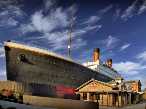 The Titanic replica stands tall beside a historic building under a blue sky with scattered clouds.