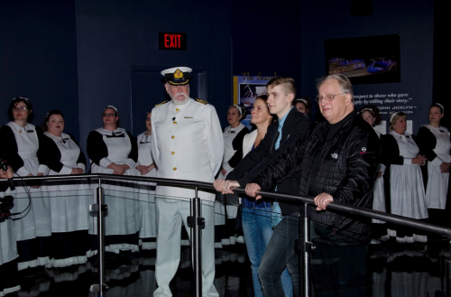 A group of people in formal attire stands in a museum, with a captain in uniform and others in historical costumes.