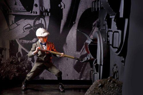 A young boy in a suit and cap shovels coal into a large black machine against a dark, industrial backdrop.