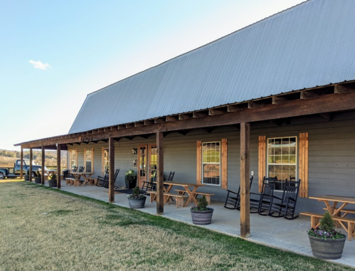 A rustic building with a metal roof, featuring a porch with rocking chairs and potted plants, set in a grassy area.