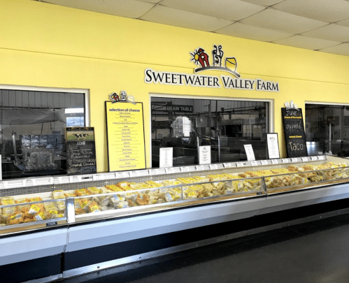 Display case filled with various cheeses at Sweetwater Valley Farm, featuring a bright yellow wall and signage.