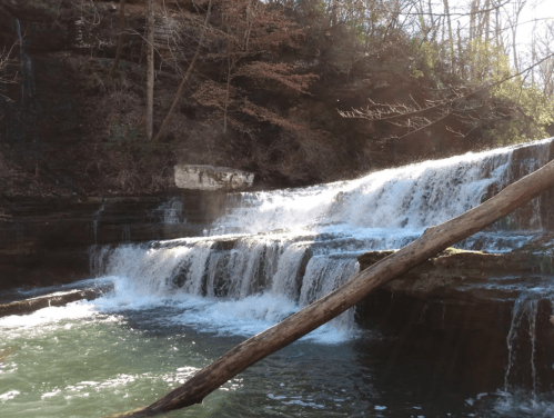 A serene waterfall cascades over rocky ledges into a clear pool, surrounded by trees and sunlight.