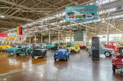 A spacious museum filled with various colorful microcars, featuring a "Microcar Mania!" banner overhead.