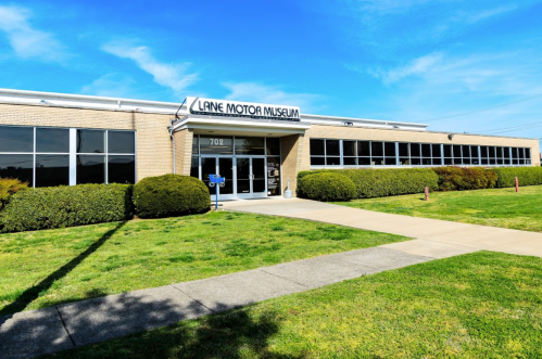 Exterior of Lane Motor Museum, featuring a brick building with large windows and a well-maintained lawn.