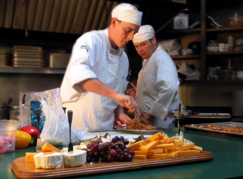 Two chefs in white uniforms prepare food in a kitchen, with a platter of cheese and grapes in the foreground.