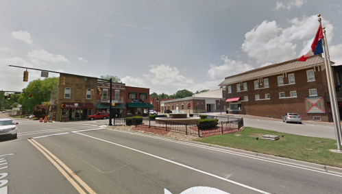 A small town intersection featuring shops, a park area, and a flagpole under a partly cloudy sky.