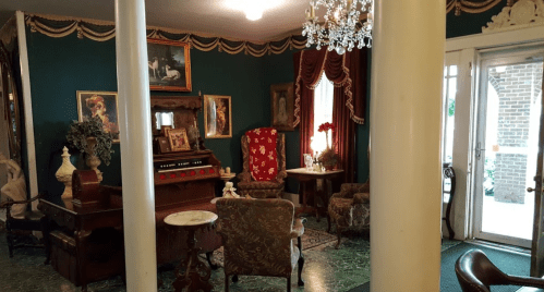 A vintage living room with ornate decor, an organ, and elegant furniture, featuring green walls and a chandelier.