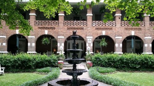 A charming brick building with arches, a fountain in the foreground, and lush greenery surrounding the area.