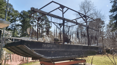 A large, old boat on a trailer, surrounded by trees and a fence, with equipment mounted above it.